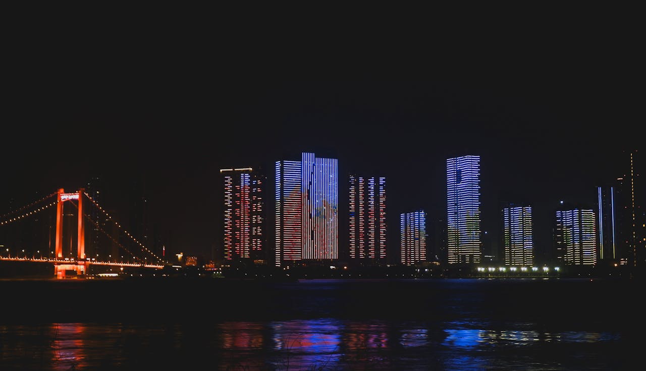 An urban nightscape featuring brightly lit high-rise buildings and a glowing bridge reflecting over a dark river.